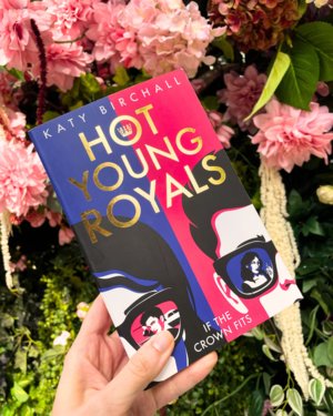 A hand holding Hot Young Royals in front of a bush of pink flowers. A hand holding Hot Young Royals in front of a bush of pink flowers.